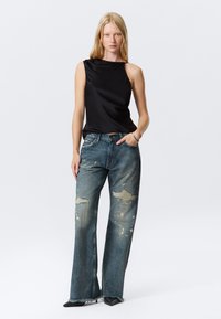 Top en satin noir avec un décolleté asymétrique, associé à un jean large bleu distressé avec des éclaboussures de peinture et des ourlets bruts.