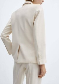Mujer vistiendo un traje color crema con una chaqueta ajustada que tiene una abertura en la parte posterior y pantalones rectos a juego.