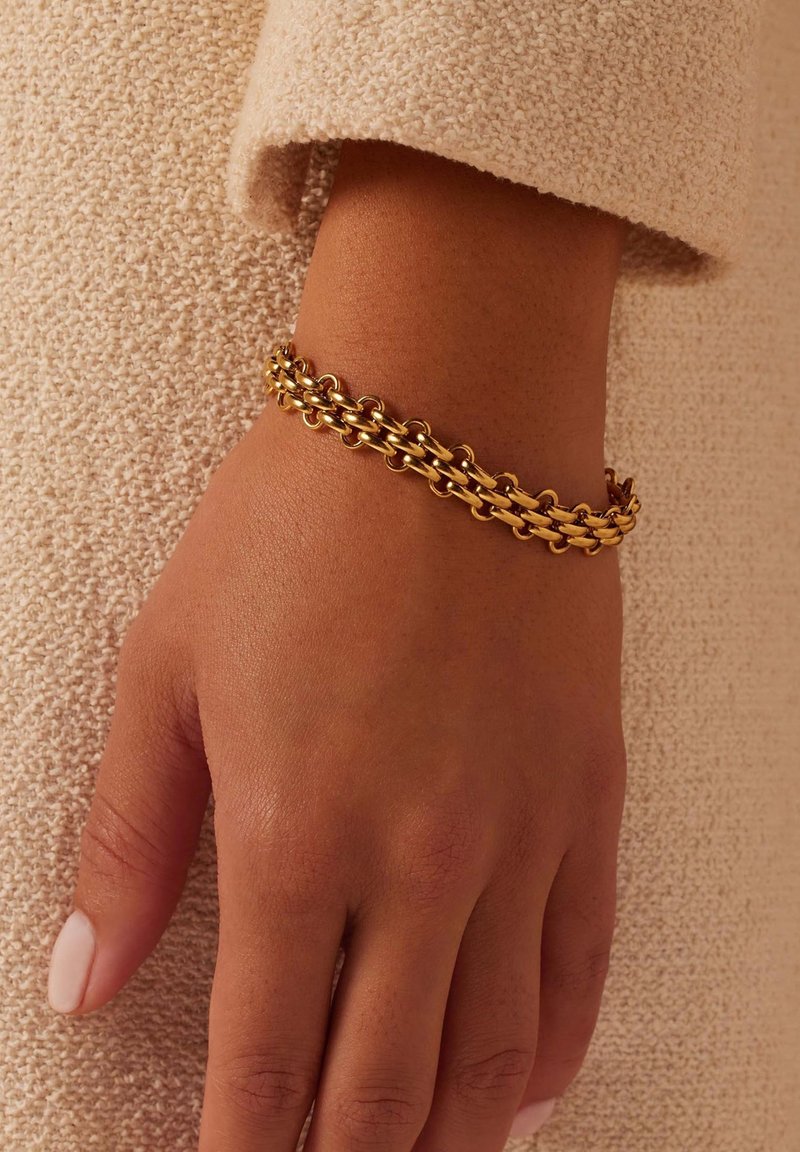 Gouden schakelarmband met elkaar verbonden ovale en ronde vormen, gepolijste afwerking, gedragen op een hand tegen een gestructureerde beige achtergrond.