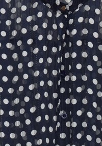 Blouse marine ornée de pois blancs, avec fermeture à boutons et texture à rayures verticales, fabriquée dans un tissu léger et transparent.