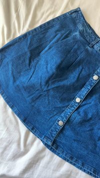 Jupe en denim bleu avec des boutons argentés sur le devant, posée à plat sur une surface en tissu blanc, montrant la texture et de légères rides.