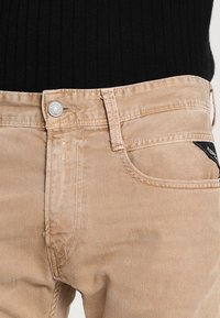 Beige cotton jeans featuring a standard five-pocket design, metal button closure, and a black label accent near the back pocket.