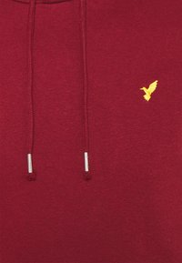 Felpa con cappuccio color bordeaux dal texture liscia, con un piccolo logo di un uccellino giallo e aghetti argentati sui lacci. Design semplice e moderno.