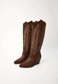 Des bottes de cowboy en daim marron présentent des orteils pointus, un talon compensé, des broderies argentées complexes et une fermeture éclair sur le côté.