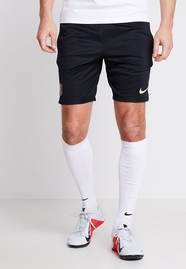 Homme portant un short de sport Nike noir avec des logos dorés, de longues chaussettes blanches et des chaussures de sport Nike blanches et rouges, debout sur fond blanc.