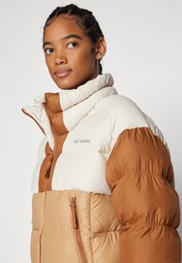 Jachetă puffer cu un design în blocuri de culoare, în nuanțe de crem și maro, cu guler �înalt, închidere cu fermoar și manșete elasticizate.