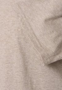 Beige tyg med synlig söm och en liten broderad logotyp nära kanten, som visar mjuk textur och fina sömnadsdetaljer.