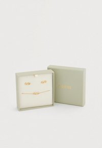 Σετ κοσμημάτων σε χρυσό τόνο σε πράσινο κουτί. Περιλαμβάνει σκουλαρίκια άπειρου και βραχιόλι, τοποθετημένα σε απαλό μπεζ μαξιλαράκι. Το όνομα της μάρκας είναι χαραγμένο σε χρυσό.