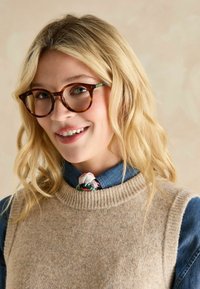 Braune Schildpattbrille mit runden Rahmen. Das Modell trägt einen beigen Strickweste über einem blauen Denimhemd, akzentuiert durch einen bunten Schal am Hals.