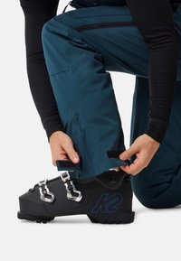 Zwarte skiboot met zilveren gespen en blauw logo; turquoise skibroek wordt met klittenband bij de enkel aangepast; getextureerde stof zichtbaar.