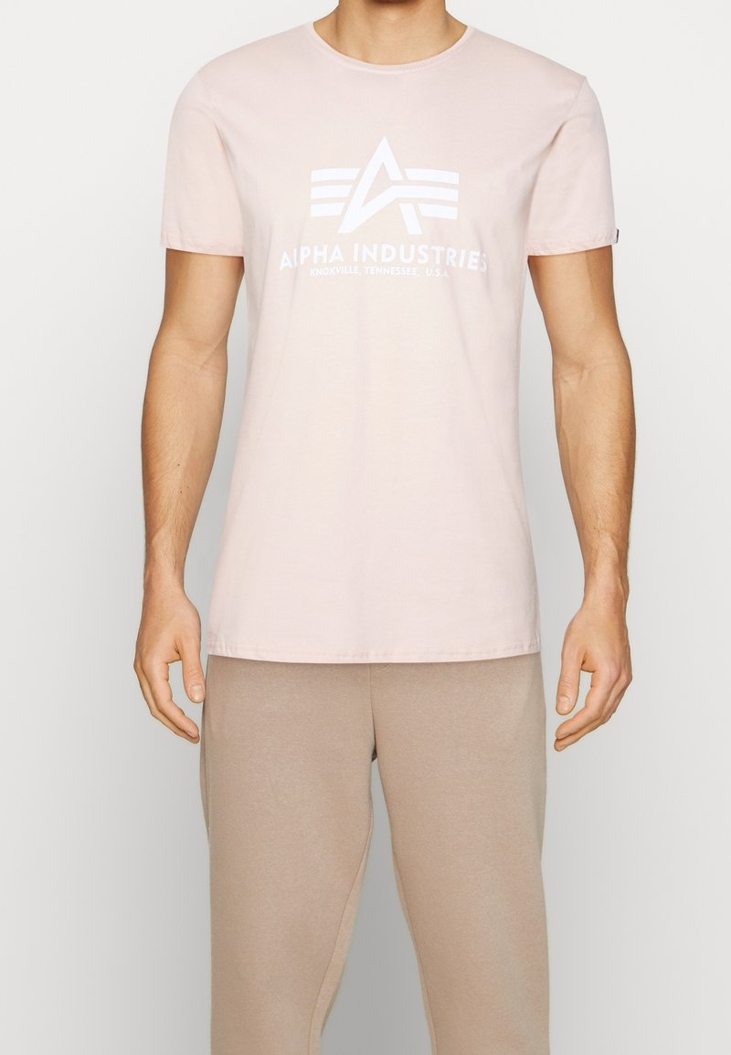 Světle růžové bavlněné tričko s krátkým rukávem a kulatým výstřihem. Na přední straně má bílé logo "ALPHA INDUSTRIES" s designovými akcenty.