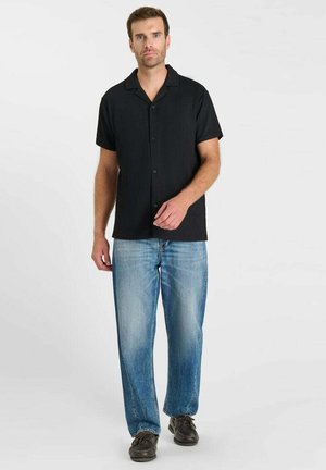 Homme debout portant une chemise noire à manches courtes boutonnée, un jean bleu clair et des chaussures marron foncé sur un fond clair uni.