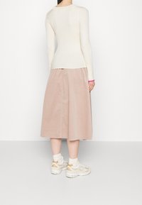 Vrouw in een crèmekleurige ribbeltrui met lange mouwen, beige midi rok, witte sokken en beige sneakers, staand met het gezicht naar een witte muur.
