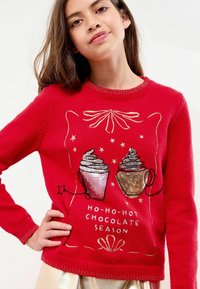 Raudonas megztinis, papuoštas dviejų puodelių karštos šokolado dizainu, puoštas blizgučiais ir žvaigždėmis, su tekstu "HO-HO-HOT CHOCOLATE SEASON."