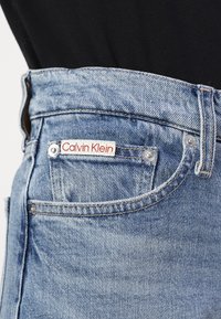 Des jeans en denim bleu clair présentent une taille haute, une étiquette Calvin Klein rouge et blanche bien visible, ainsi que des rivets en métal argenté sur les poches.