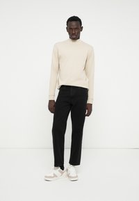 Maglietta beige a maniche lunghe abbinata a jeans neri, con una vestibilità dritta e sneakers bianche con dettagli beige e superficie testurizzata.