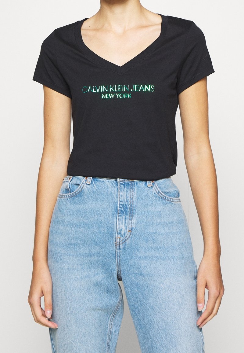 Black cotton T-shirt with a V-neck, featuring reflective "CALVIN KLEIN JEANS NEW YORK" text. Paired with light blue high-waisted jeans.
