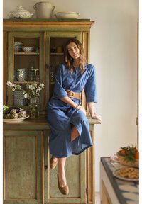 Žena v modrých pruhovaných šatech sedí na rustikální zelené dřevěné skříni s květinami, nádobím a zeleninou v jemně osvětlené místnosti.