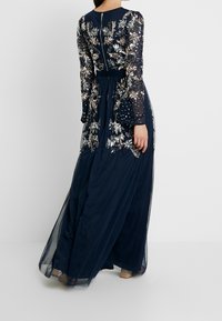 Marinblå långklänning med genomskinliga, dekorerade ärmar, blommig broderi i silver och guld, och en svävande, transparent kjol.