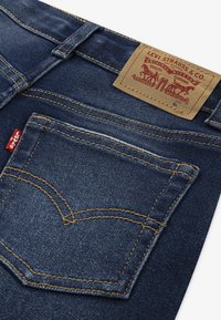 Mørkeblå denimjeans med en synlig baklomme med et gult stikningsmønster og et merket lærpatch på midjebåndet.