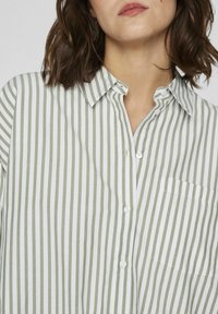 Camisa de rayas en blanco y verde salvia, diseño de botones, con cuello, un solo bolsillo en el pecho y una textura suave.