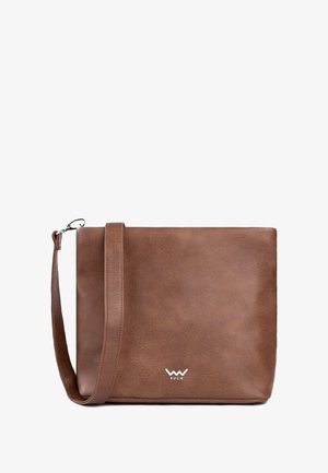 Gesteppte beige Weste, braunes Oberteil, braune Leggings und weiße Stiefeletten. Braune Schultertasche. Holzhütte und Steg in natürlicher Umgebung.