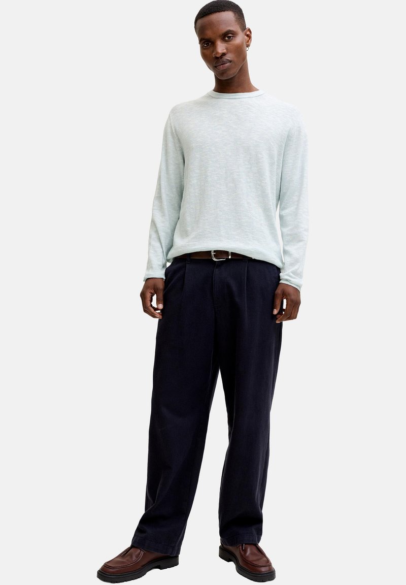 Homme portant une chemise à manches longues gris clair, un pantalon noir ample avec ceinture, et des chaussures habillées marron, debout devant un fond uni.