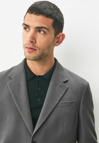 Grau strukturiertes Blazer mit strukturierten Revers, kombiniert mit einem schwarzen Poloshirt mit drei Knöpfen. Sauberes Design mit Fronttaschendetail.
