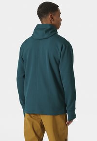 Suéter de punto en color verde azulado con cuello alto y mangas largas. Presenta un patrón texturizado y un corte relajado, combinado con pantalones amarillos.