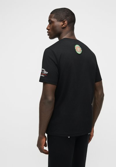 Homme tourné vers la gauche portant un t-shirt noir avec un écusson rond sur le haut du dos et un texte blanc-rouge-vert sur la manche droite.