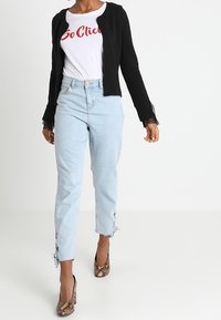 Zwarte cardigan met kantaccenten, witte t-shirt met rode tekst, lichtblauwe jeans met rafelige zoom en gestreepte hoge hakken.