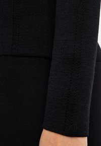 Nahaufnahme eines schwarzen gestrickten Ärmels und einer Taille mit strukturierten vertikalen Mustern, der am Arm und Oberkörper einer Person getragen wird.