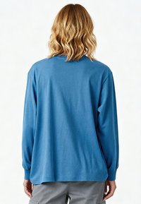 Personne aux cheveux blonds ondulés mi-longs portant une chemise bleue à manches longues et un pantalon gris, vue de dos.
