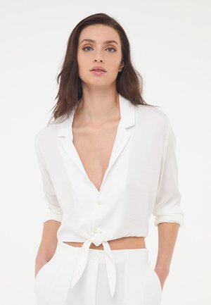Camicia bianca, leggera e corta, con un nodo in vita e maniche arrotolate, caratterizzata da un profondo scollo e una texture liscia.