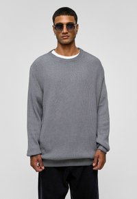 Grijze gebreide trui met een losse pasvorm, ronde halslijn en ribbelstructuur, gedragen met donkere broek en ronde zonnebril.