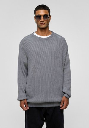 Grijze gebreide trui met een losse pasvorm, ronde halslijn en ribbelstructuur, gedragen met donkere broek en ronde zonnebril.