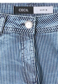 Gros plan sur un pantalon en denim rayé verticalement bleu et blanc, avec un bouton argenté et une étiquette CECIL DENIM à la ceinture.