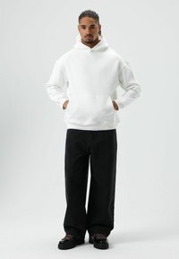 Witte hoodie met capuchon en voorzak, gecombineerd met losvallende zwarte broek en zwarte schoenen met contrasterende veters.