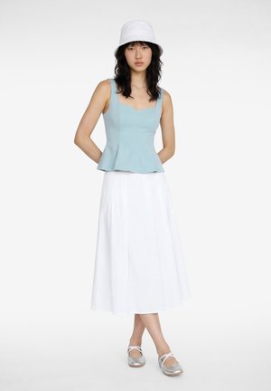 Ung kvinde iført en hvid bucket hat, lyseblå ærmeløs top, hvid midi nederdel og sølv ballet flats, stående med armene bag ryggen.