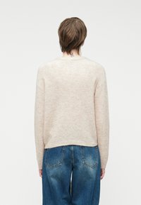 Helle beige Wollpullover mit weicher Textur, Rundhalsausschnitt und abgehängten Schultern. Getragen mit lockeren blauen Denim-Jeans.