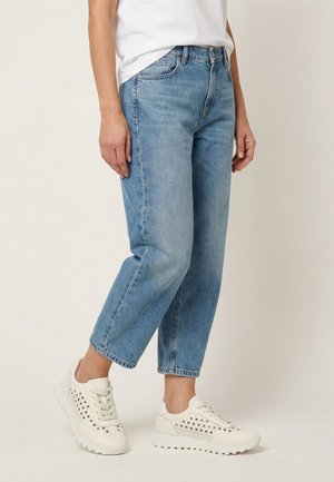 Persoon die lichtblauwe cropped jeans, witte geperforeerde sneakers en een wit T-shirt draagt, staand op een effen lichte achtergrond.