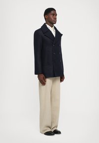 Námornícky vlnový pakoat s dvojradovým zapínaním na gombíky a bočnými vreckami, spárovaný s krémovými širokými nohavicami a čiernymi topánkami.