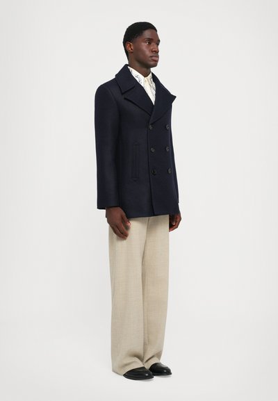 Jūras spēku zila vilnas jūreivju mētelis ar divus rindu pogām un sānu kabatām, apvienots ar krēmkrāsas platām biksēm un melnām kurpēm.