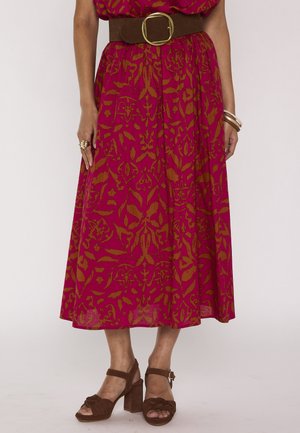 Une femme porte une jupe midi à motifs magenta et marron, une large ceinture marron avec une grande boucle dorée, des sandales marron à talons hauts et des bracelets.