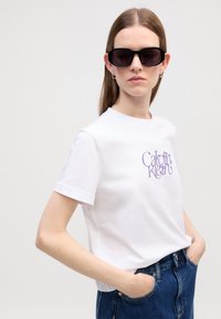 T-shirt en coton blanc avec un design de logo "Calvin Klein" violet. Porté avec un jean en denim bleu foncé taille haute et des lunettes de soleil noires rectangulaires.