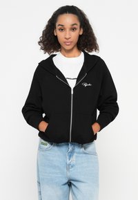 Crna rebrasta zip-up dukserica s velikim kapuljačom, prednjim džepovima i bijelim izvezenim logom. Uparena s laganim širokim trapericama.