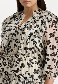 Blommig klänning i genomskinligt tyg med svarta bladmönster, volang runt halsen och puffärmar. Ljus bas med rynkad midja.