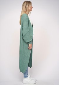 Dlouhý pletený kardigan v světle zelené barvě s volným střihem, který má oversized rukávy a otevřený přední design. Doplňený džíny a teniskami.