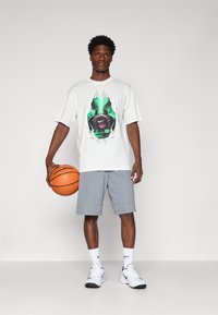 Hvid grafisk t-shirt med et farverigt print i midten, grå træningsshorts, hvide sokker og sort-hvide sneakers. Holder på en basketball.