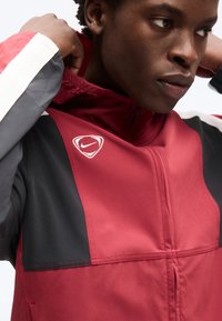 Νεαρό άτομο που προσαρμόζει το γιακά ενός αθλητικού μπουφάν Nike σε κόκκινο και μαύρο χρώμα με λευκό λογότυπο στο στήθος.